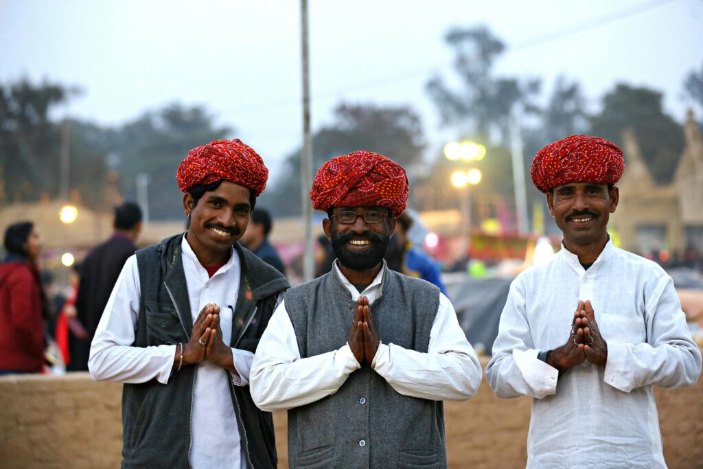 Three Indian men wearing traditional clothing and turbans smiling outdoors during the day.